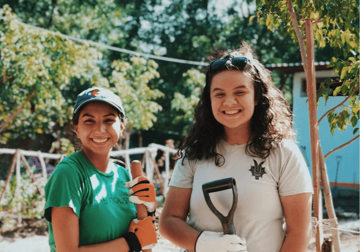 Two young women smile warmly at the camera while volunteering outdoors on a sunny day. Both wear work gloves and hold gardening tools — one in a green t-shirt and baseball cap holds a tool handle, while the other in a light gray t-shirt with sunglasses propped on her head holds a shovel.